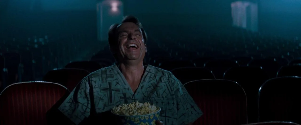 John Trent laughing alone in an empty movie theater with a bucket of popcorn